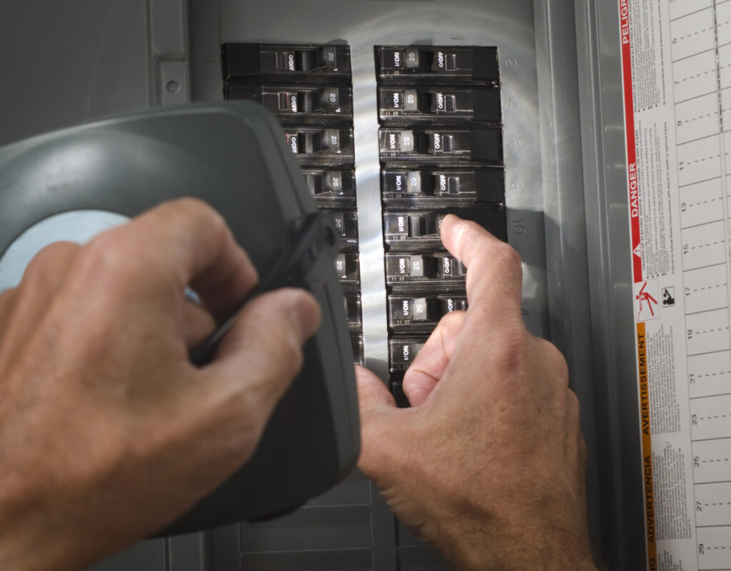 Technician's hands holding flashlight while flipping switch on electrical panel.