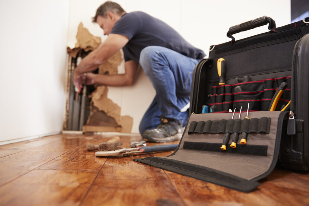 Plumbing repairing burst pipe behind a wall in a home, focus on tool bag in foreground