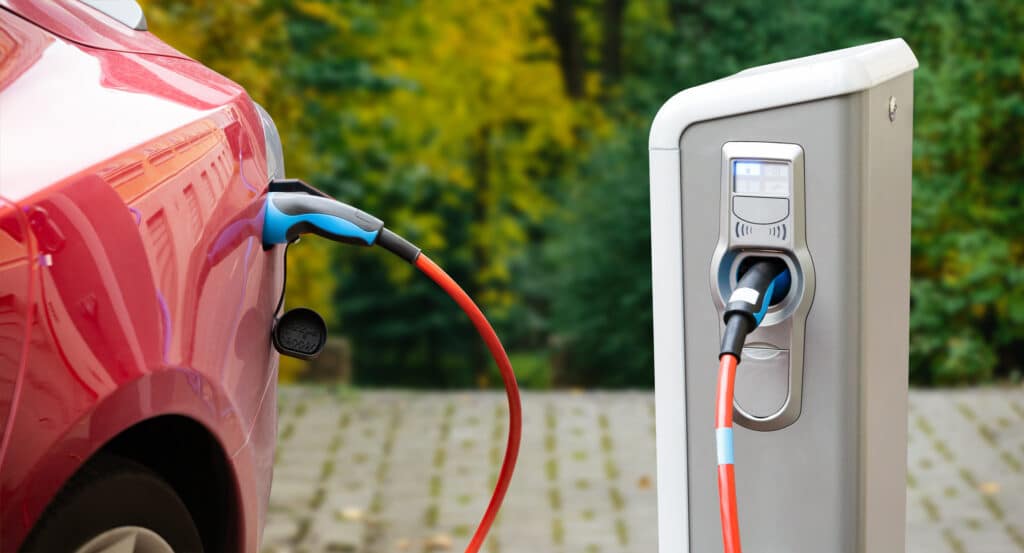 Close-up view of a red electric car plugged into an electric vehicle charging station outside.