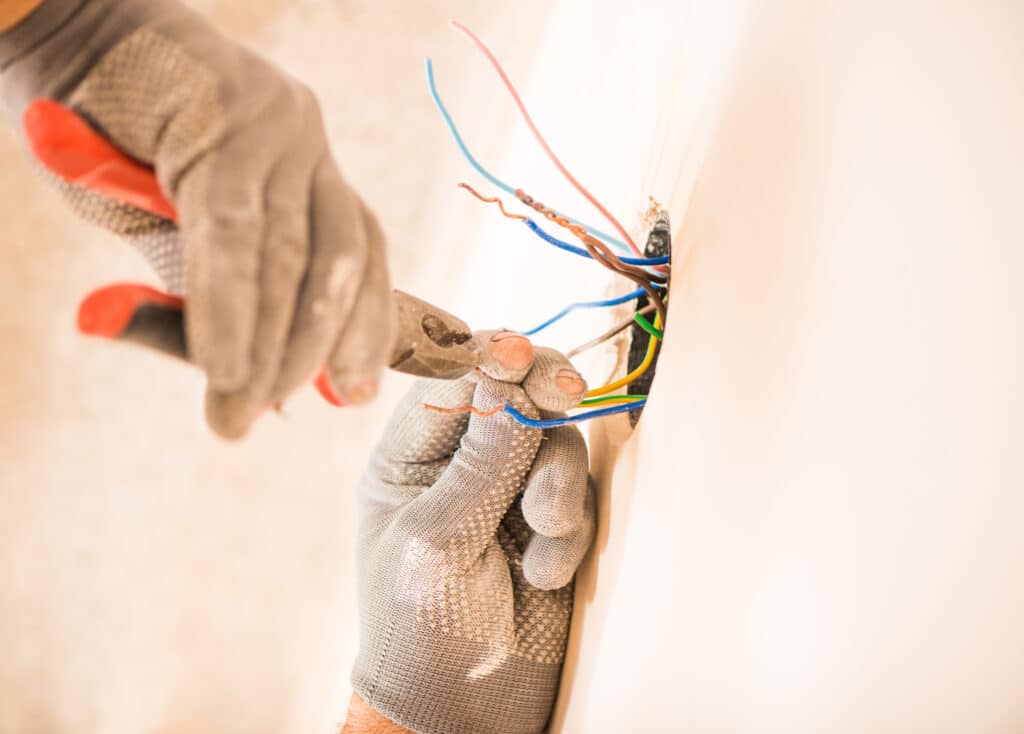 Technician in gloves using pliers working with electrical wires through a hole in a white wall.