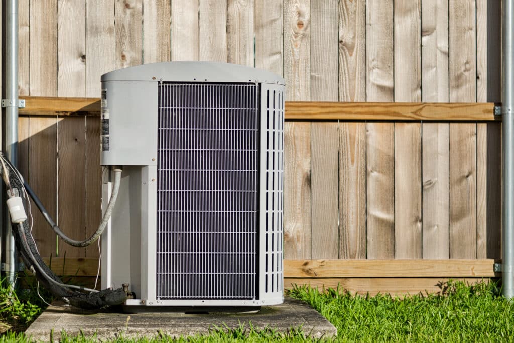 Central air conditioning unit in a residential backyard in front of a wooden fence and lawn area.