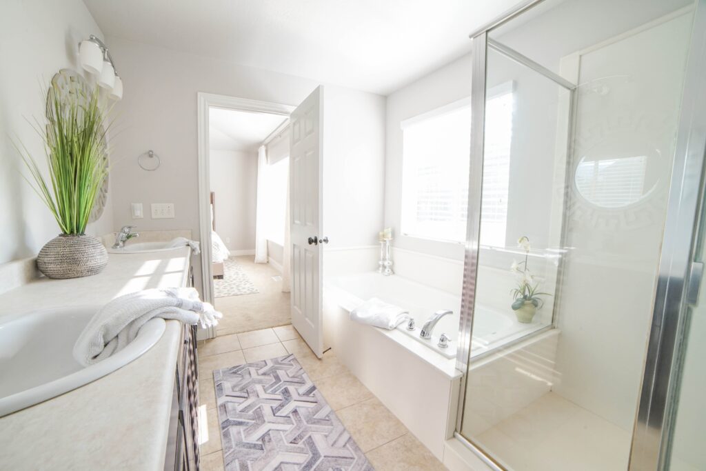 Residential bathroom complete with a vanity, separate tub, toilet, and walk-in shower. White walls, large window, and light tan tile floors.