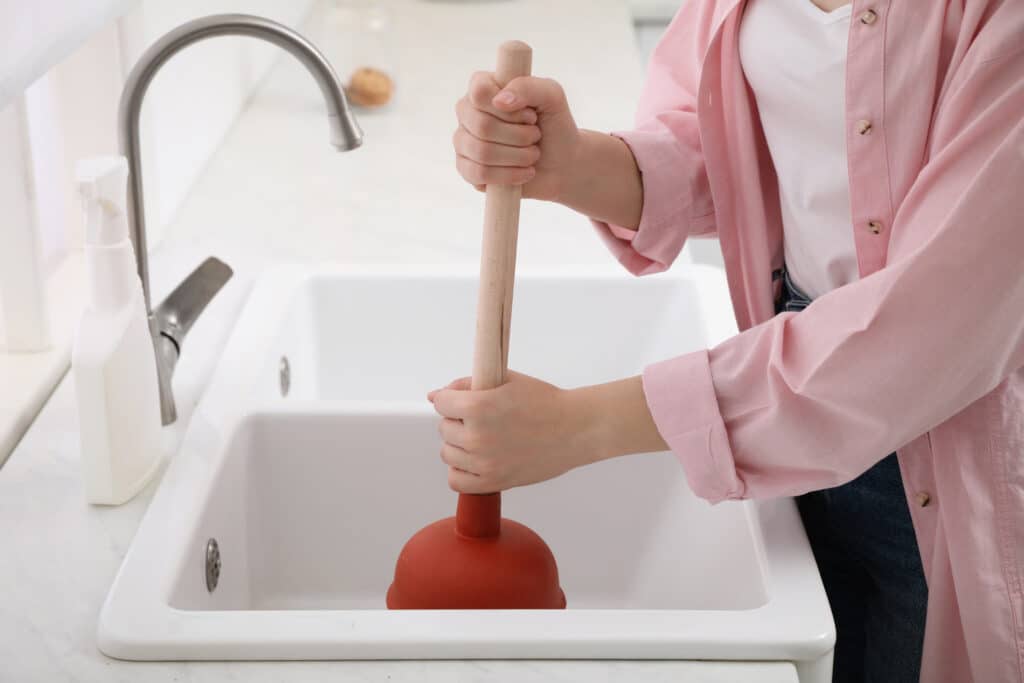 Woman using plunger to unclog sink drain in kitchen.