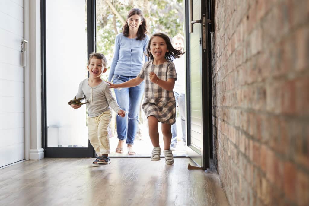 Excited children running through front door of home with their mother behind them, smiling.