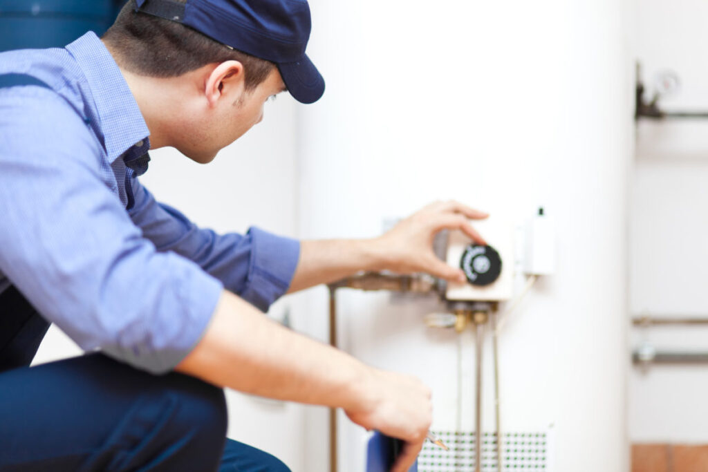 Plumber kneeling next to white tank water heater and adjusting a knob.