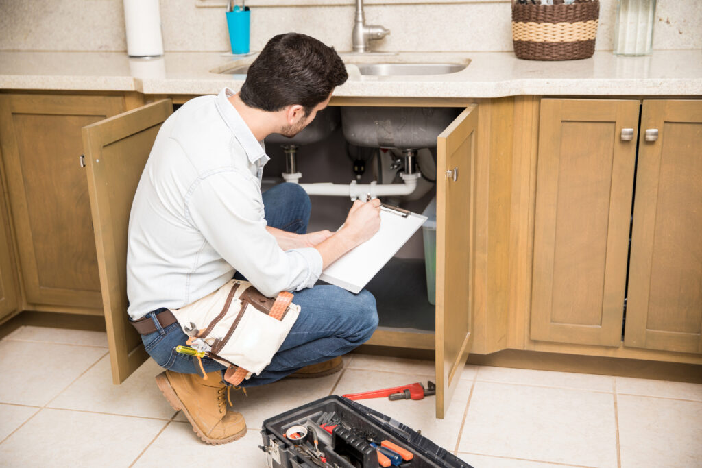 Plumber writing a repair order while crouching in front of a kitchen sink with cabinet open underneath.