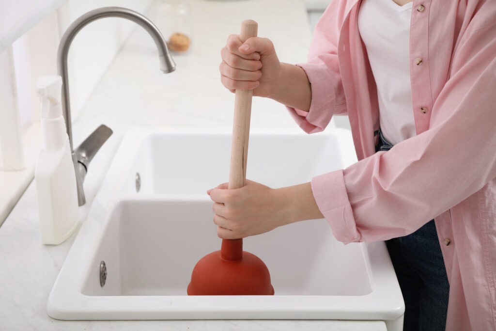 Woman wearing light pink shirt, using plunger to unclog sink drain in kitchen.